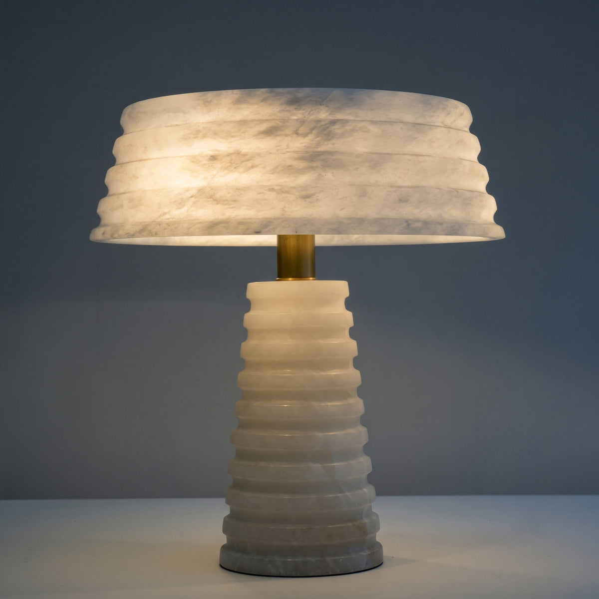 Orivon 3 Light Sculpted Alabaster Table Lamp in soft white, with a ribbed tapered base and banded drum shade joined by a brass finial, lit against a plain grey background.