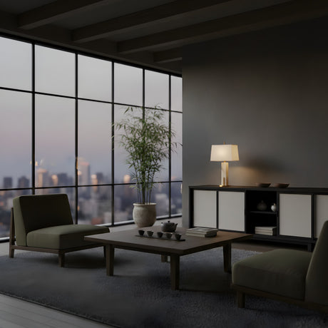 A modern living room with floor-to-ceiling windows and minimalist decor features a matte black coffee table, a potted plant, and the Strataform 1 Light Framed Alabaster Table Lamp in Matte Black & Soft White glowing softly on the sideboard.