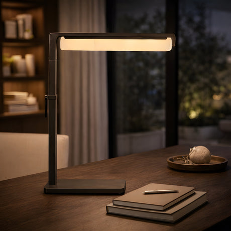 The Linea LED Alabaster Table Lamp in Matte Black glows on a wooden desk beside closed books, a pen, and a decorative tray. Shelves and an evening-lit window set a calm, contemporary scene.