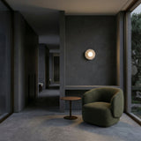 A modern, minimalist room with dark gray walls, large windows, a Caldera 1 Light Small Disc Travertine Wall Light in light beige, a green armchair, and a round wooden side table. Natural and ambient lighting enhance the serene interior.