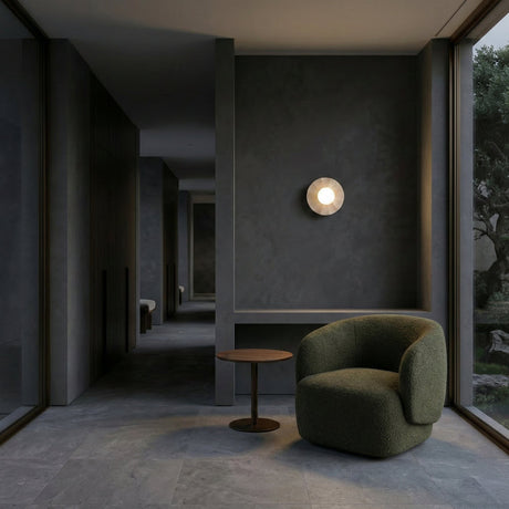 A modern, minimalist room with dark gray walls, large windows, a Caldera 1 Light Small Disc Travertine Wall Light in light beige, a green armchair, and a round wooden side table. Natural and ambient lighting enhance the serene interior.