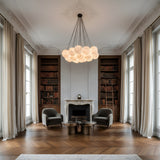 Elegant library with tall windows, floor-to-ceiling bookcases, a white ornate fireplace, two modern armchairs, round coffee tables, parquet floors, and the Auriel 19 Light Alabaster Chandelier in Soft White & Black hanging from a decorative ceiling.