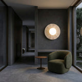 A modern, minimalist interior with dark walls and floors features the Caldera 1 Light Large Disc Travertine Wall Light in light beige, a green curved armchair, and a small round side table near large windows overlooking greenery.