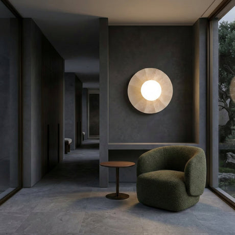 A modern, minimalist interior with dark walls and floors features the Caldera 1 Light Large Disc Travertine Wall Light in light beige, a green curved armchair, and a small round side table near large windows overlooking greenery.