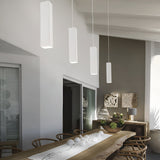 In this contemporary dining room, a long wooden table is complemented by charming chairs. Four Emberlux 1 Light Pendants in white hang above, casting a warm glow. The sideboard displays decorative items, while artfully stacked logs adorn the wall. Sunlight filters through shutters, enhancing the serene ambiance.
