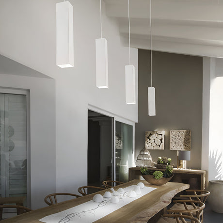 In this contemporary dining room, a long wooden table is complemented by charming chairs. Four Emberlux 1 Light Pendants in white hang above, casting a warm glow. The sideboard displays decorative items, while artfully stacked logs adorn the wall. Sunlight filters through shutters, enhancing the serene ambiance.