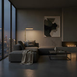 A modern living room at dusk features city views, a gray sectional sofa, abstract wall art, and a minimal coffee table. The Linea LED Alabaster Floor Lamp in brushed brass casts a warm glow over the inviting space.