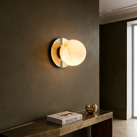 The Noctyra 1 Light Alabaster Wall Light in Soft White & Brass is mounted on a dark green wall above a wooden console table holding a “Brancusi” book and gold decor, with natural light streaming through a nearby window.