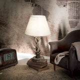 A cozy vintage-style room features a weathered armchair with a soft throw blanket. The Flow Wood Table Lamp - Dark Brown, with its nature-inspired design and white shade, sits atop books beside an antique radio, all set against a wallpapered wall adorned with old newspaper prints.