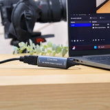 AV:Link HDVC4Kv2 4K HDMI Video Capture Card connected between an HDMI cable and a laptop running capture software on a wooden desk.