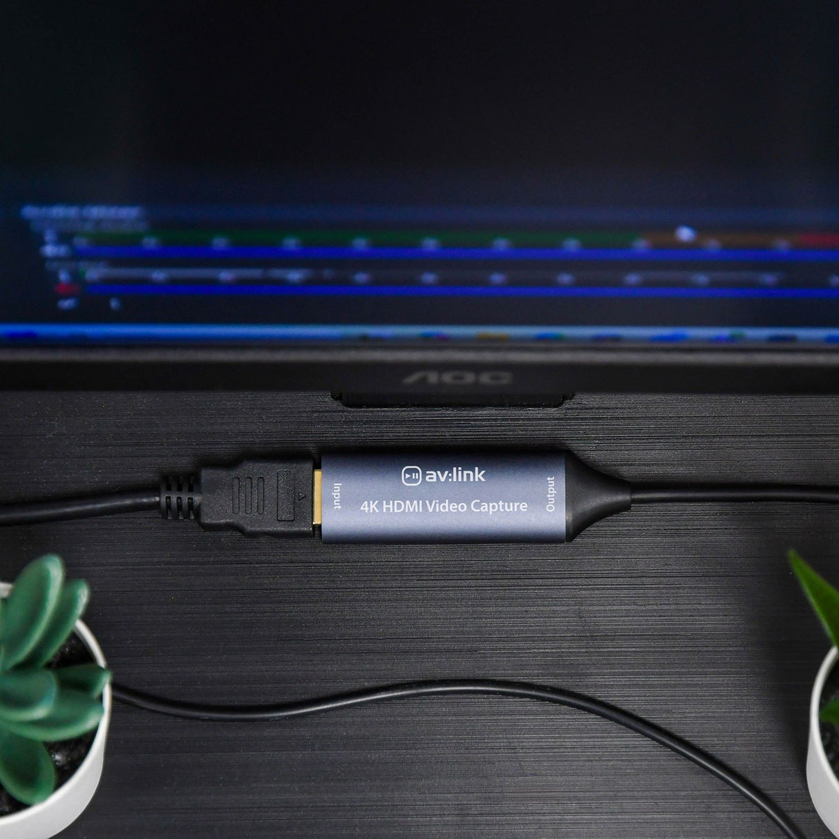 AV:Link HDVC4Kv2 4K HDMI Video Capture Card connected to a monitor on a dark desk, with HDMI input and USB output cables between two small plants.