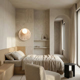 A minimalist bedroom with neutral tones features a wooden bed, light bedding, the Serano 1 Light Disc Travertine Wall Light in light beige, built-in shelves with decor, a soft armchair, a small black side table, and floor-length curtains by large windows.