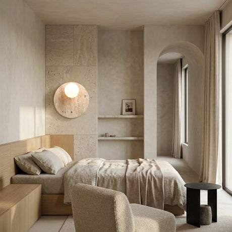 A minimalist bedroom with neutral tones features a wooden bed, light bedding, the Serano 1 Light Disc Travertine Wall Light in light beige, built-in shelves with decor, a soft armchair, a small black side table, and floor-length curtains by large windows.