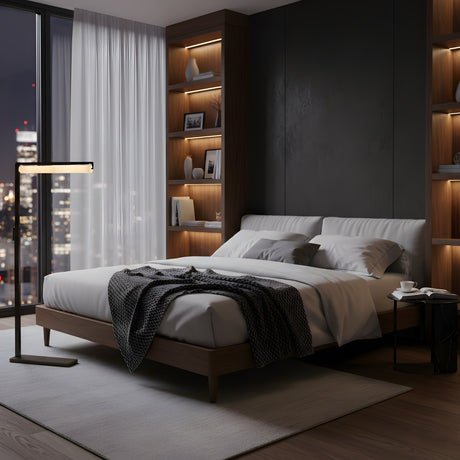 A modern bedroom featuring a large bed with gray bedding, a dark knitted throw, built-in wooden shelves with decor, and the Linea LED Alabaster Floor Lamp in Matte Black by the window overlooking city skyline views at night.