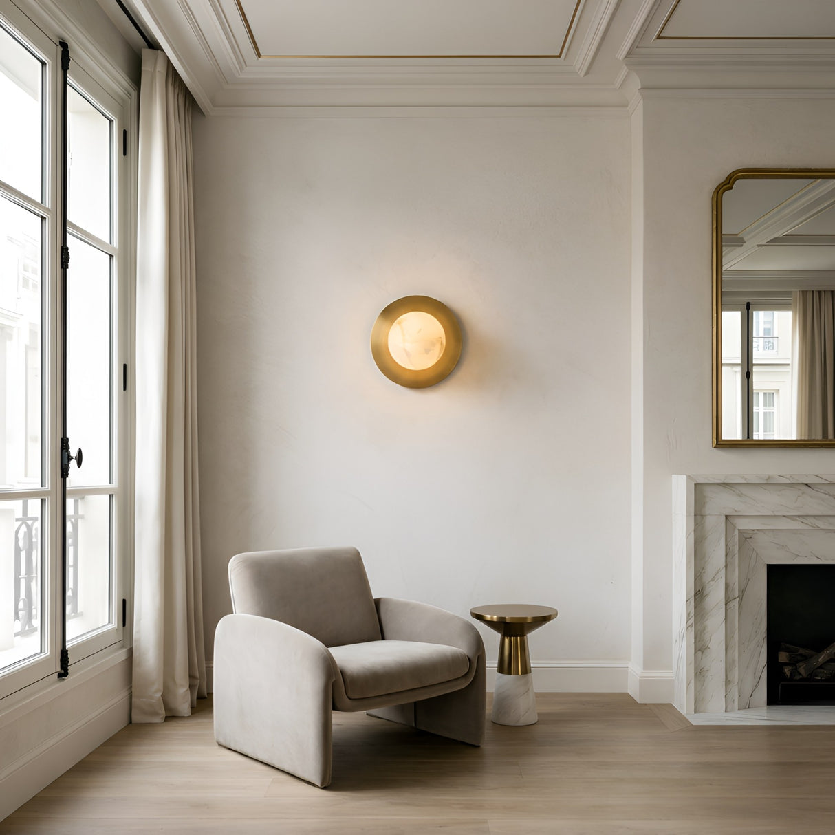 A modern, minimal living room featuring a beige armchair, round accent table, marble fireplace, large curtained window, gold-framed mirror, and the Noctyra 1 Light Alabaster Wall Light in soft white & brass on a white wall.