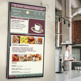 AV:Link fixed vertical anti-theft TV wall bracket shown installed in portrait, holding a digital menu display on a wall inside a café setting.