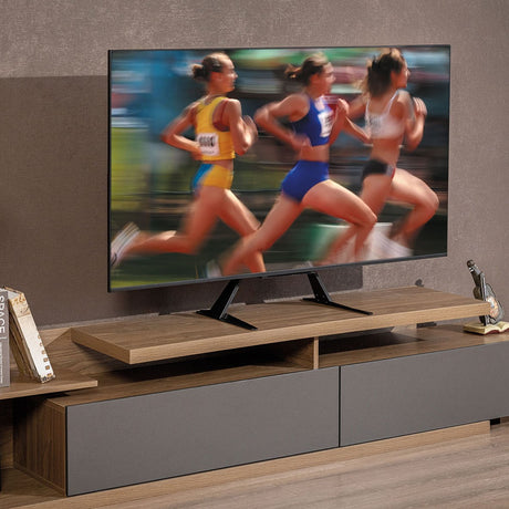Large flat-screen TV mounted on a black AV:Link PS800 tabletop stand, set on a wood-tone TV unit in a living room.