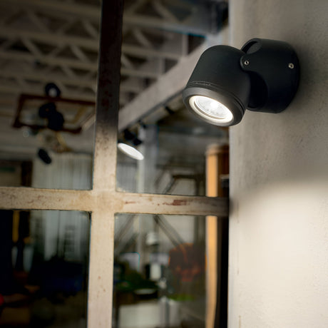 The Vista Outdoor Wall Spotlight - Black is illuminated next to a window, casting light on the rustic interior with exposed beams and glass. This wall-mounted spotlight enhances the modern industrial design and boasts an IP44 rating for durability in varied conditions.