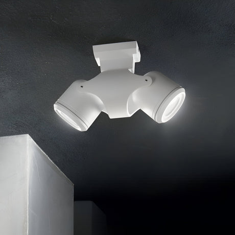 The Vista Outdoor Twin Spotlight - White, a contemporary ceiling light fixture made from powder-coated aluminium, showcases two adjustable round LED lamps. When installed on a dark ceiling, it directs focused beams downward, providing partial illumination to an adjacent wall.