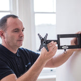 Man in a navy polo shirt adjusting the AV:Link HQT201 full motion double arm TV wall bracket in black steel against a white interior wall.