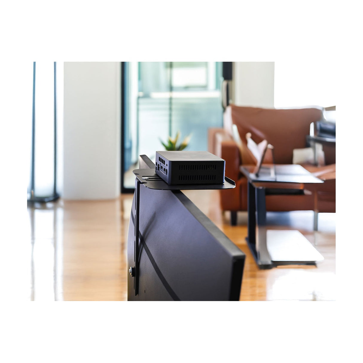 AV:Link ACS300 TV accessory shelf mounted behind a flat-screen TV holding a small black media box, shown in a bright living room setting.