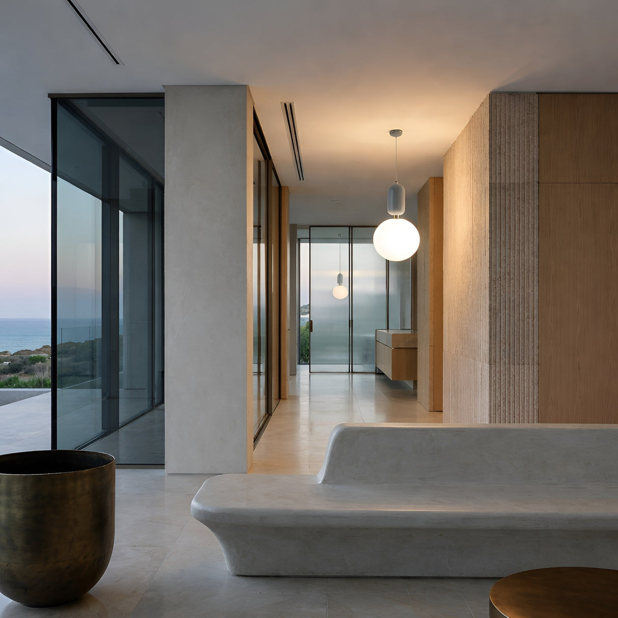 A modern minimalist interior with large glass windows, beige walls, a sculpted stone bench, the Pallo Pendant Light - White with an opal glass diffuser, and a brass cylindrical planter—overlooking sea views at sunset.