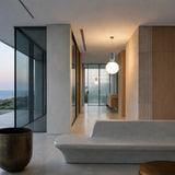 A modern minimalist interior with large glass windows, beige walls, a sculpted stone bench, the Pallo Pendant Light - White with an opal glass diffuser, and a brass cylindrical planter—overlooking sea views at sunset.