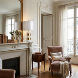 Elegant living room with ornate white walls, a large mirror above a white fireplace, vintage books and vases on the mantel, a plush armchair with pillow and throw, and the Arvella 1 Light Striped Marble Table Lamp in White & Gold by tall, sheer-curtained windows.