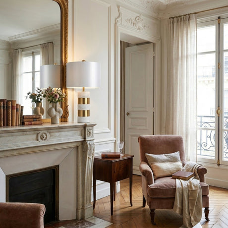 Elegant living room with ornate white walls, a large mirror above a white fireplace, vintage books and vases on the mantel, a plush armchair with pillow and throw, and the Arvella 1 Light Striped Marble Table Lamp in White & Gold by tall, sheer-curtained windows.