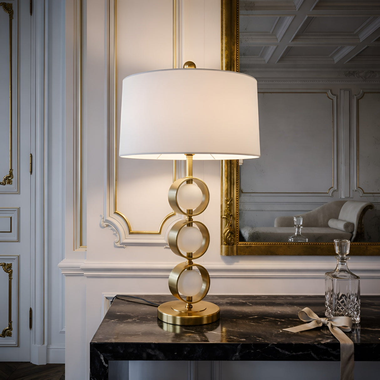 The Wisp 1 Light Stacked Orb Marble Table Lamp - Brass & White with a white shade sits on a black marble console table in an elegant room with ornate white and gold paneling, alongside a glass decanter tied with a ribbon and a tumbler.