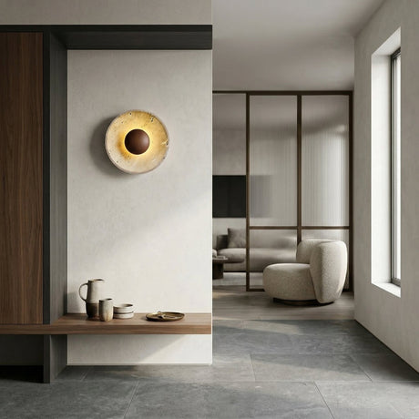 A minimalist interior in neutral tones features a Torello LED Round Travertine Wall Light in Walnut Brown & Light Beige, a wooden bench with ceramics, gray floor tiles, and a cozy chair in a sunlit living area with glass-paneled dividers.