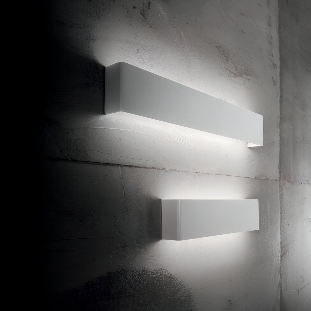 The Canvas LED Wall Light 23W 3000K - White, each measuring 60cm in length, casts a soft, ambient glow against the textured gray wall. Featuring a sleek and minimalist design, these energy-efficient lights are installed vertically atop one another to create a modern and stylish ambiance.