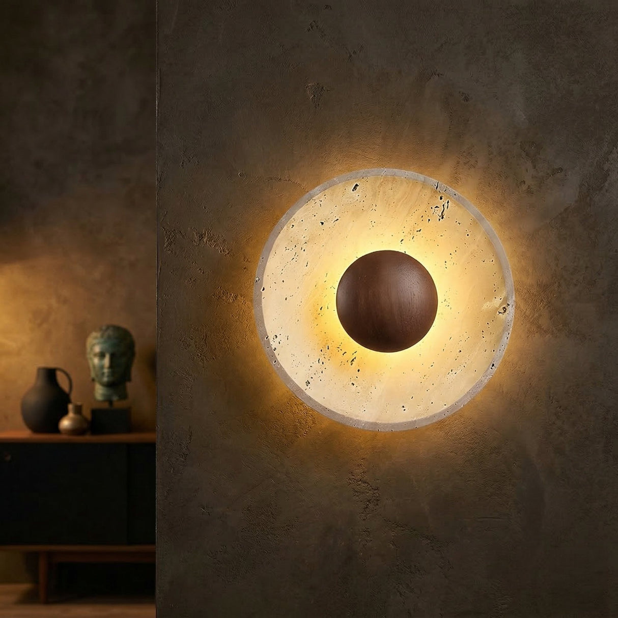 The Torello LED Round Travertine Wall Light in Walnut Brown & Light Beige, with its glowing circular frame and dark center, is mounted on a textured walnut brown wall; a sideboard behind features a vase, sculpture, and decor items.