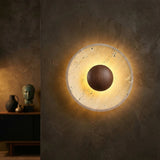 The Torello LED Round Travertine Wall Light in Walnut Brown & Light Beige, with its glowing circular frame and dark center, is mounted on a textured walnut brown wall; a sideboard behind features a vase, sculpture, and decor items.