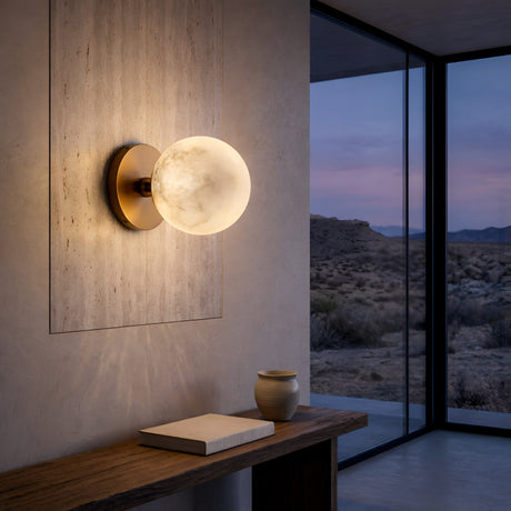 The Lunavere 1 Light Sphere Alabaster Wall Light in soft white and brass glows warmly on a textured wall above a wooden table with a book and ceramic vase, as desert views stretch beyond large glass windows at sunset.