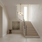 A modern staircase with wooden steps and a black handrail leads to a bright window. Above, the Astra 5 Light Alabaster Pendant Light in Soft White & Brushed Brass hangs, while light walls and floor enhance the airy space.
