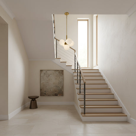 A modern staircase with wooden steps and a black handrail leads to a bright window. Above, the Astra 5 Light Alabaster Pendant Light in Soft White & Brushed Brass hangs, while light walls and floor enhance the airy space.