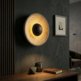 The Torello LED Round Travertine Wall Light in Walnut Brown & Light Beige emits a warm glow against a dark textured wall above a desk with decor, while a framed abstract art piece complements the stylish, dimly lit room.