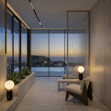 Modern minimalist room with two low chairs, a Pallo Table Lamp - Black, and a planter by floor-to-ceiling windows framing a serene sunset. Neutral tones and sleek design, elevated by minimalist lighting, create a calm, inviting atmosphere.