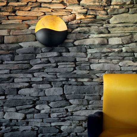 The Beamrise 2 Light Wall Light in gold brightens up a rustic stone wall with its modern design. In the foreground, the vivid yellow of an armchair stands out against the textured gray stones, offering ambient lighting that cultivates a cozy and welcoming environment.