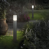 As dusk settles over the garden, two Wave Outdoor Bollard Lights in grey, designed to withstand the elements, illuminate a stone path bordered by lush greenery and flower beds, enhancing the tranquil ambiance of the setting.