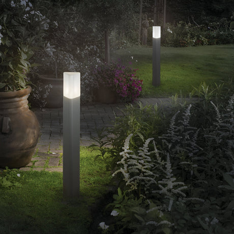As dusk settles over the garden, two Wave Outdoor Bollard Lights in grey, designed to withstand the elements, illuminate a stone path bordered by lush greenery and flower beds, enhancing the tranquil ambiance of the setting.