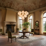 Niori Astra 13 light alabaster cluster chandelier in brushed brass hangs above a round table in a frescoed Tuscan-style room with arched windows.