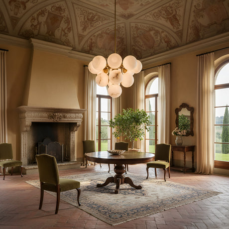 Elegant room with ornate ceiling, arched windows, stone fireplace, round wooden table, four green chairs, potted plant, decorative rug, classic furnishings, and the Astra 12 Light Alabaster Pendant Light in soft white & brushed brass. Sunlight streams in.