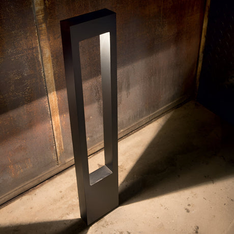 The Phasia Outdoor Pedestal Light in black features a modern, rectangular design with an open, geometric structure that illuminates a textured, rustic wall and concrete floor. Its sleek black finish contrasts beautifully with the warm tones of its surroundings, making it perfect for outdoor lighting thanks to its IP44 protection rating.