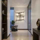 A modern, minimalist hallway featuring light wood floors, the Mireya 4 Light Metal Chain Veil Wall Light in Nickel, a gray chair by a large window with cityscape views, a potted plant, and dark wood furniture with chic decor accents.