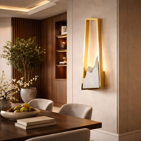 A modern dining room with a wooden table, light gray chairs, and a fruit bowl. The wall features the Atria LED Left Alabaster Wall Light – Brushed Brass. Built-in shelves, décor objects, and a green plant decorate the background.