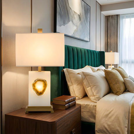A stylish bedroom features a green upholstered bed, beige bedding with gold pillows, and a wooden nightstand with books and the Selune 1 Light Sculpted Core Alabaster Table Lamp in Brushed Brass & Soft White. Another lamp and window are seen in back.