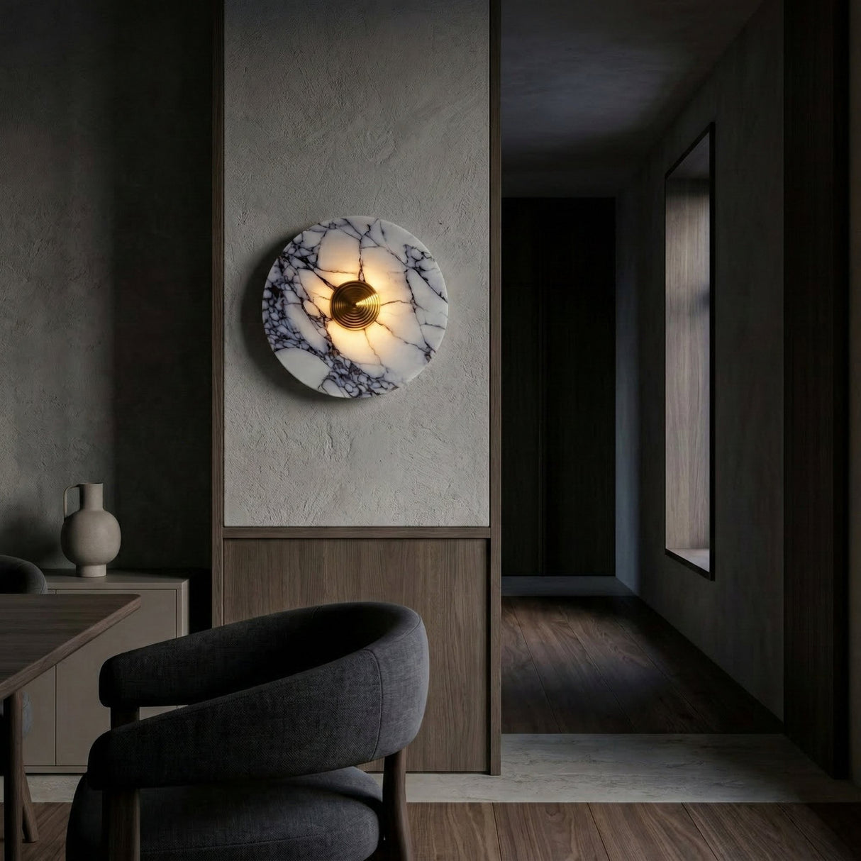 A modern dining room features the Carrara LED Small Veined Disc Marble Wall Light in white, black, and brushed brass, paired with dark upholstered chairs, a wooden table, and minimalist decor that complements the hallway’s wooden flooring.
