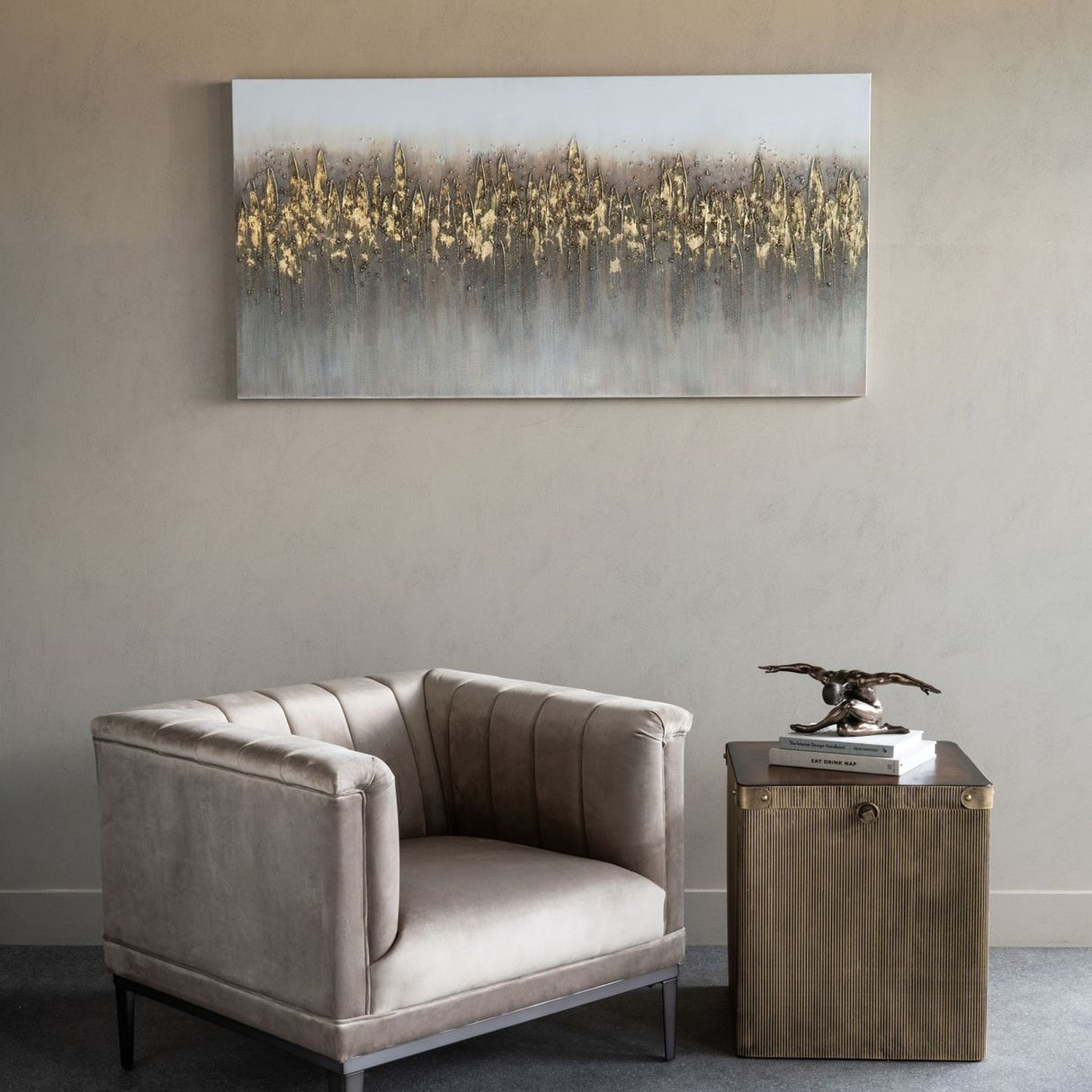 Libra Interiors Bronze Male Arms Outstretched Sculpture on books atop a ribbed side table, beside a beige velvet armchair and abstract gold artwork.